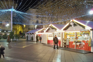 Ein lebendiger Weihnachtsmarkt in der Nacht in einer Stadt, mit Menschen, die stehen, sitzen und eine Person, die ein Kind hält, umgeben von festlichen Lichtern, Pfählen, Pflanzen, Bäumen und Gebäuden mit Fenstern unter einem bewölkten Himmel.