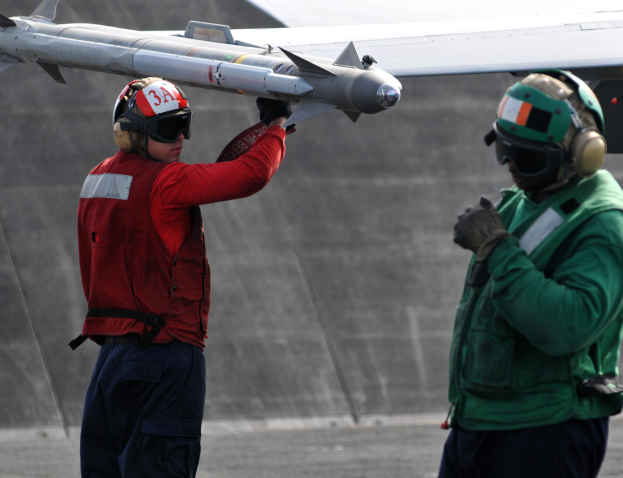 Zwei M├Ąnner in Helmen und Handschuhen stehen auf einer Rollbahn, einer h├Ąlt eine Rakete, waehrend der andere sie betrachtet, mit einem Flugzeug im Hintergrund.