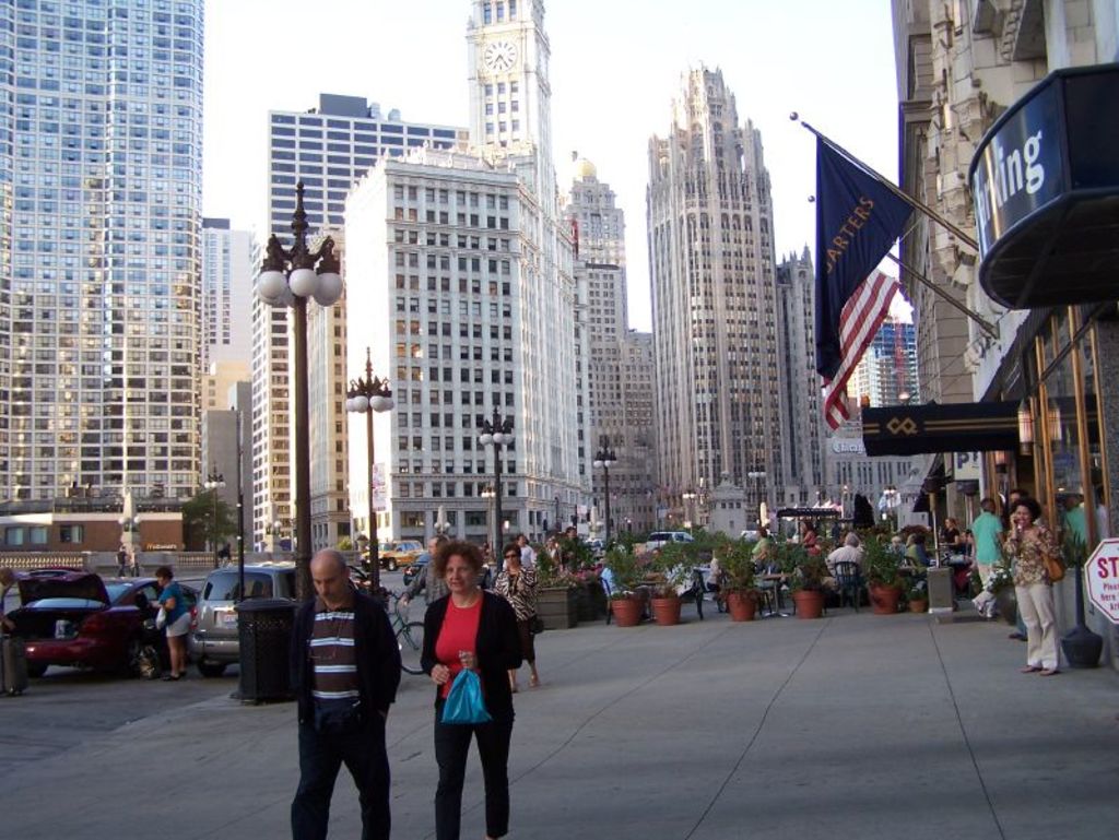 Eine belebte Straßenszene mit Fußgängern, Blumentöpfen, Autos, Straßenlaternen, Gebäuden im Hintergrund, Geschäften auf der rechten Seite und Fahnen.