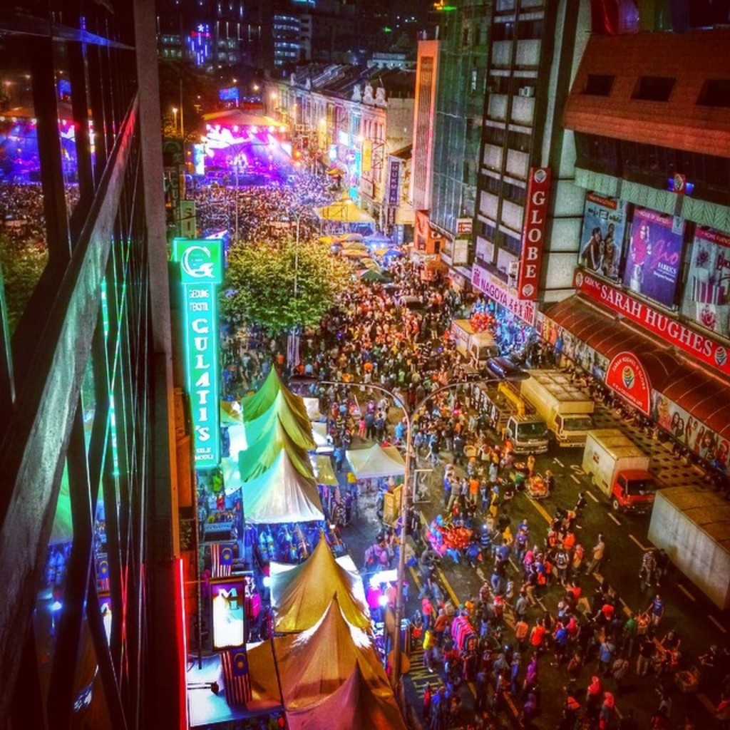 Eine lebhafte Karnevalsstimmung mit einer großen Menschenmenge auf der Straße, umgeben von Zelten, Geschäften, Gebäuden und Bannern, mit zahlreichen Lichtern, die den Hintergrundbereich erhellen.