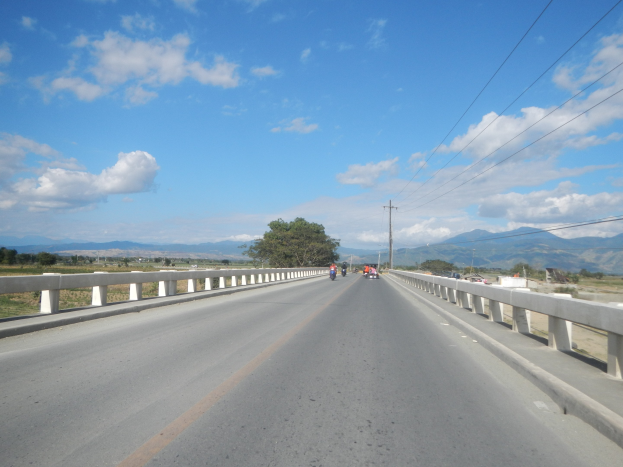 Autobahn mit Motorradfahrern, Geländern, Strommasten, Bäumen, Hügeln und bewölktem Himmel im Hintergrund.