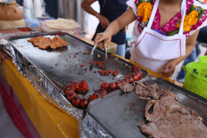 Eine Frau in einer Schürze grillt auf einem Markt, mit einem Tisch voller Lebensmittel, einem Eimer und anderen Gegenständen in der Nähe und ein paar Menschen im Hintergrund.