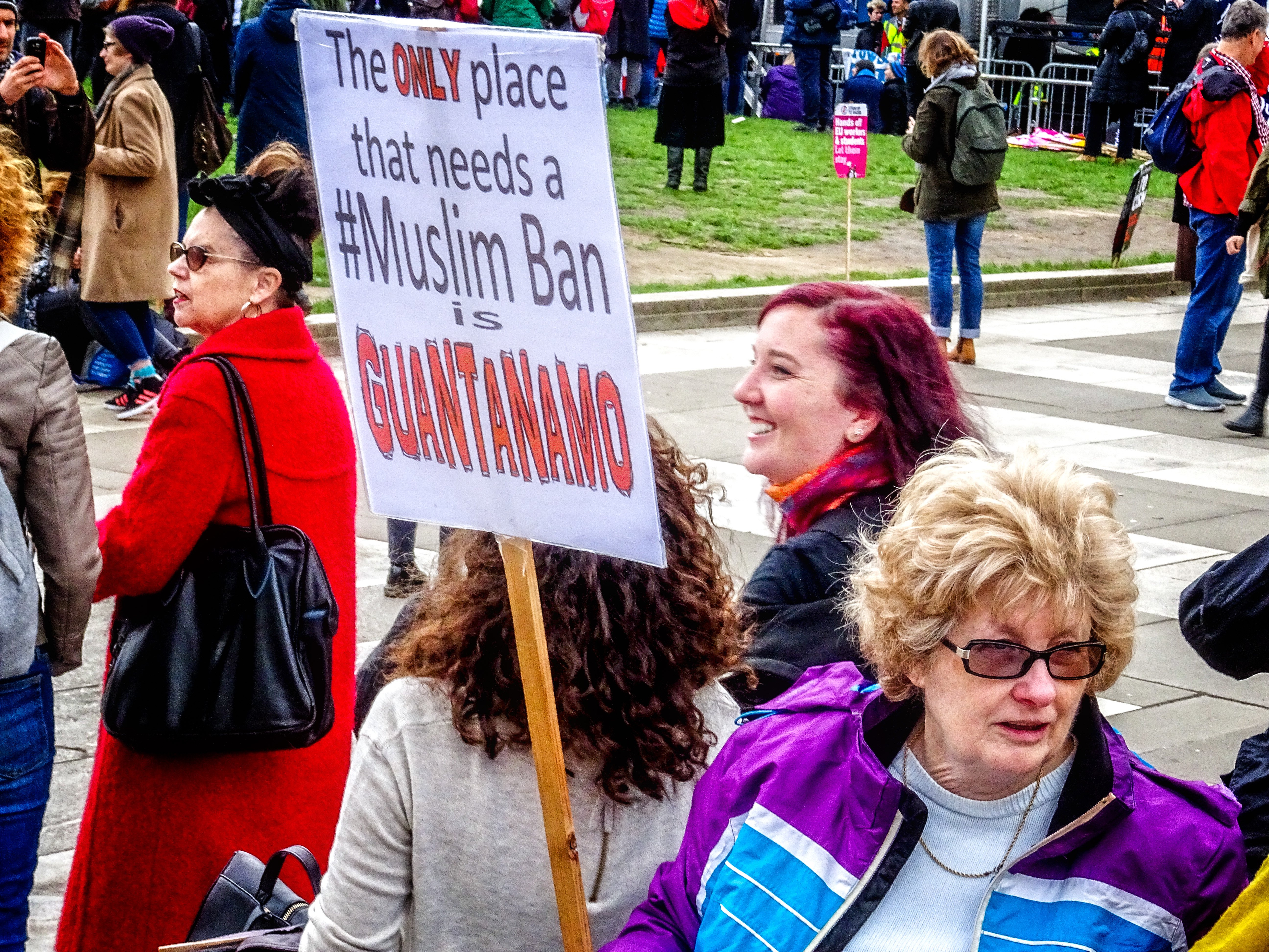 Eine Gruppe von Menschen steht auf dem Boden, einige halten Taschen, eine Frau hält ein Schild mit der Aufschrift "Der einzige Ort, an dem ein Muslimverbot notwendig ist, ist Guantanamo". Im Hintergrund sehen wir einen Metallzaun, Gras, ein Schild mit Text, einige Pfosten, ein Gebäude mit Fenstern und Bäume.