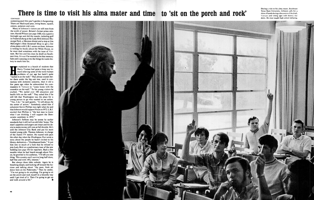 Eine Person mit Brille steht vor einer Gruppe sitzender Personen, mit einer Wand mit Vorhängen und Fenstern im Hintergrund.