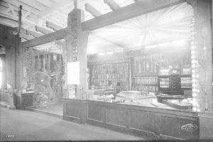 Ein altes Schwarz-Weiß-Foto einer Apotheke mit einem Tresen, der verschiedene Gegenstände zeigt, Regalen mit Flaschen und anderen Gegenständen im Hintergrund und Text am unteren Rand, der darauf hinweist, dass es sich um eine Apotheke handelt.