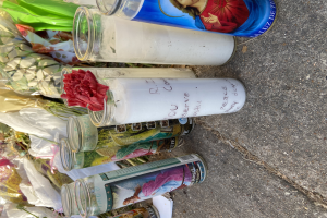 Eine improvisierte Gedenkstätte am Straßenrand mit Glasflaschen, die Fotos und Text zeigen, einem Blumenstrauß und brennenden Kerzen auf dem Boden.