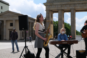 Eine Frau spielt Saxophon vor dem Brandenburger Tor in Berlin, Deutschland, mit einer Person, die ein Piano spielt, einer anderen, die eine Gitarre hält, und einem Lautsprecher im Vordergrund, während im Hintergrund Gebäude, Bäume und Wolken zu sehen sind.