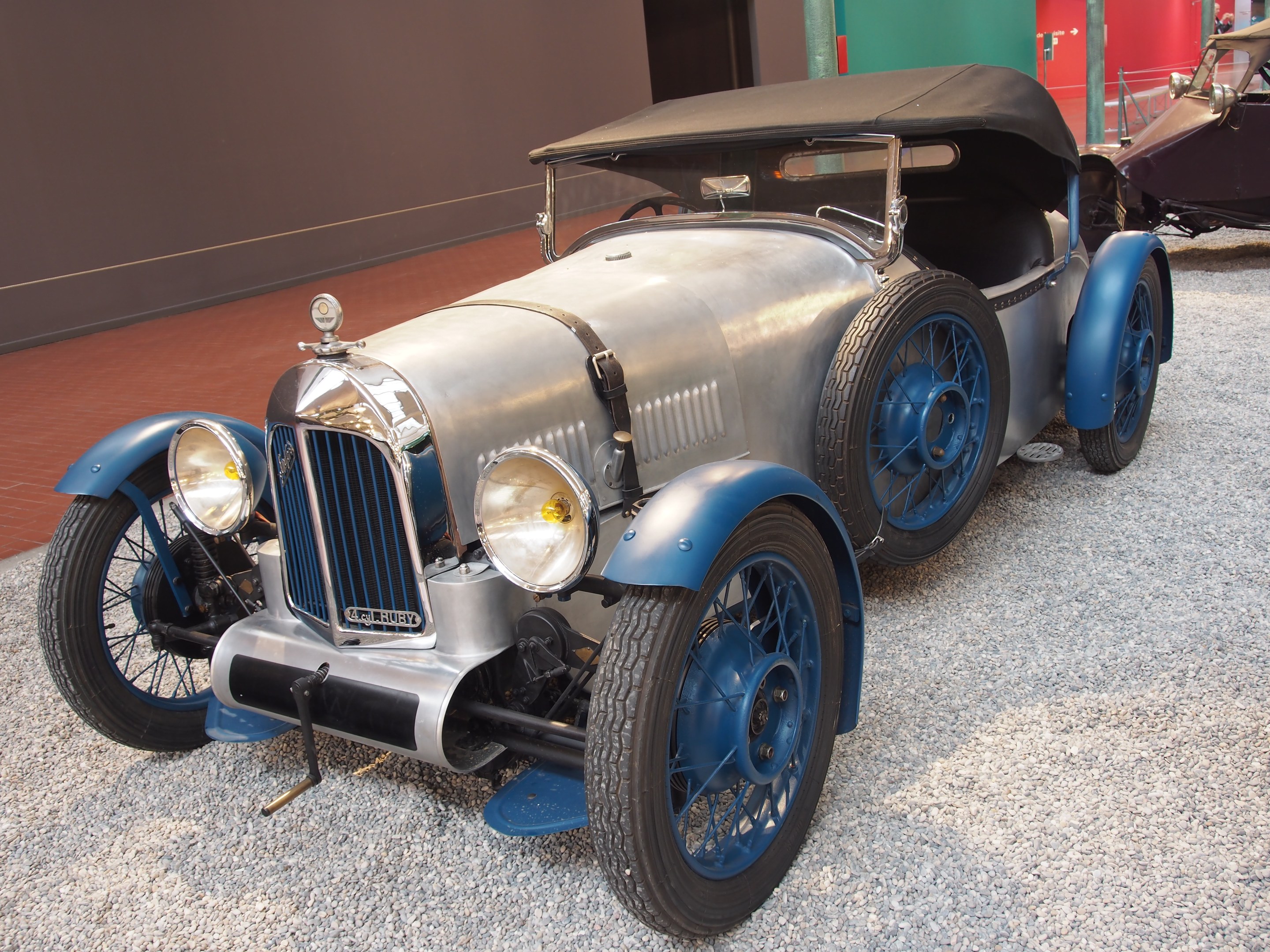 Ein Oldtimer-Rumbo-Sitz-Auto mit glänzendem Finish in einem Museum ausgestellt, umgeben von anderen Fahrzeugen, mit einer Wand, einer Tür und Menschen im Hintergrund.