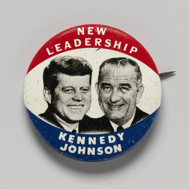 Ein Ansteckbutton auf einer weißen Oberfläche zeigt zwei Fotos von Kennedy und Johnson mit dem Text 'Neue Führung' darunter.