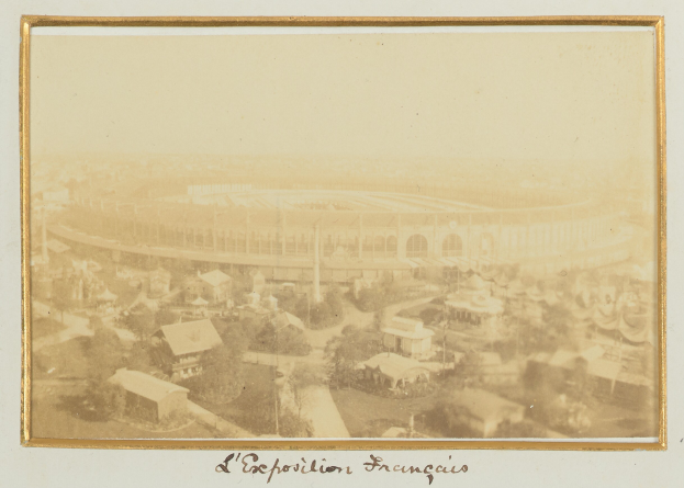 Schwarz-weiß-Foto eines Stadions umgeben von Gebäuden, Bäumen und Strommasten mit Text unten.