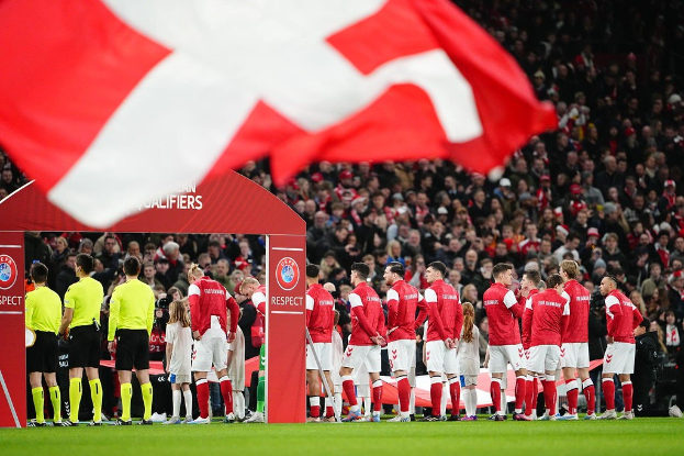Eine Gruppe von Menschen auf einem Fußballfeld mit einer roten und weißen Flagge im Vordergrund, einem Bogen mit dem Text 'Bayern München vs. Bayern München Wetten & Vorschau' im Hintergrund und einer großen Menschenmenge im Stadion.