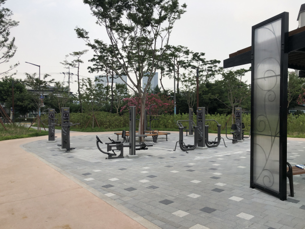 Freiluftpark mit verschiedenen Fitnessgeräten, Bänken, Bäumen, Pflanzen, Gras, Masten, Lichter, Kabel und Gebäuden unter dem Himmel.