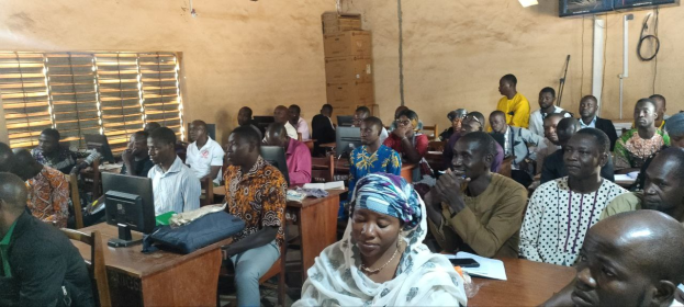 A group of people seated at desks in a classroom with monitors, bags, books, and papers, attending a technology training session.