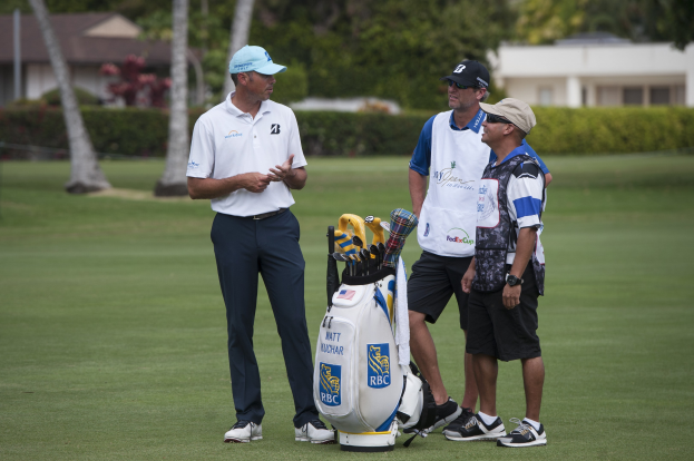 Mannergruppe auf grünem Feld mit Golfbag und -clubs im Vordergrund, Bäume und Gebäude im Hintergrund.