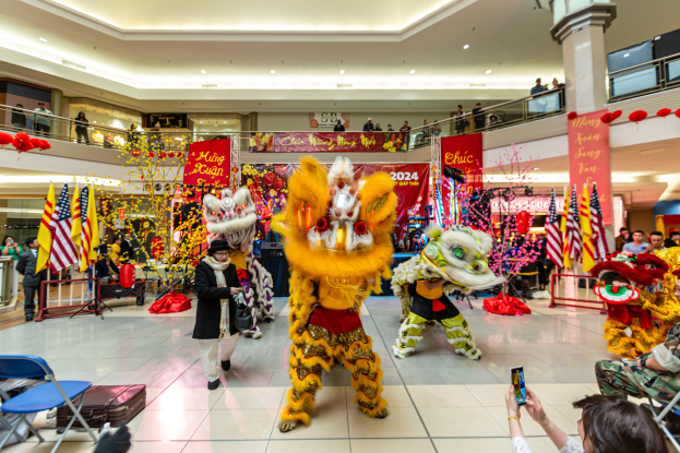 Ein lebhaftes chinesisches Neujahrsfest in einem Einkaufszentrum mit tanzenden Menschen in Kostümen, anderen auf Stühlen sitzend mit Mobiltelefonen und verschiedenen Dekorationen, Flaggen und Bannern im Hintergrund.