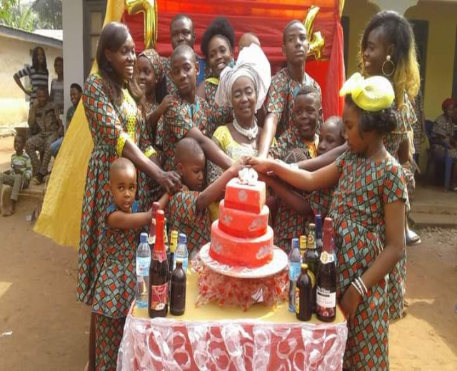 Eine Gruppe von Menschen sitzt um einen Tisch mit einer Torte, Flaschen und anderen Gegenständen in einer ländlichen Umgebung mit Häusern, Bäumen und einem klaren blauen Himmel, was auf eine Geburtstagsfeier hindeutet.