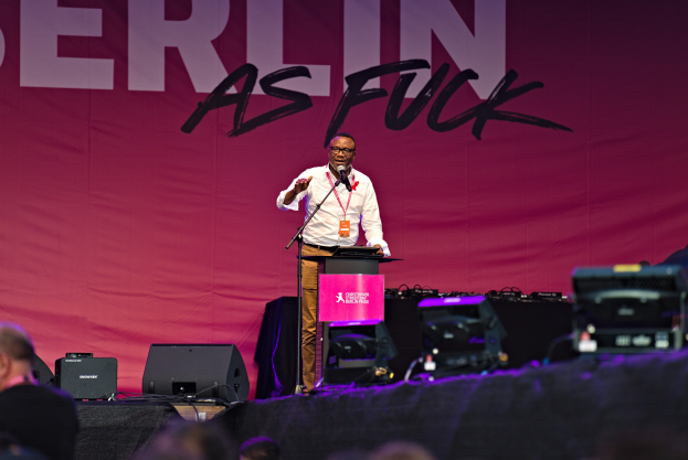 Mann in weißem Hemd und braunen Hosen, der in ein Mikrofon auf einer Bühne spricht, mit einem pinken "Berlin as Fuck"-Plakat im Hintergrund.