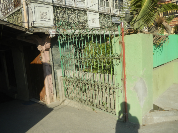 Ein altes Gebäude mit einem grünen, kunstvoll gestalteten Metalltor, umgeben von Bäumen und Pflanzen, mit mehreren Fenstern und Balkonen.