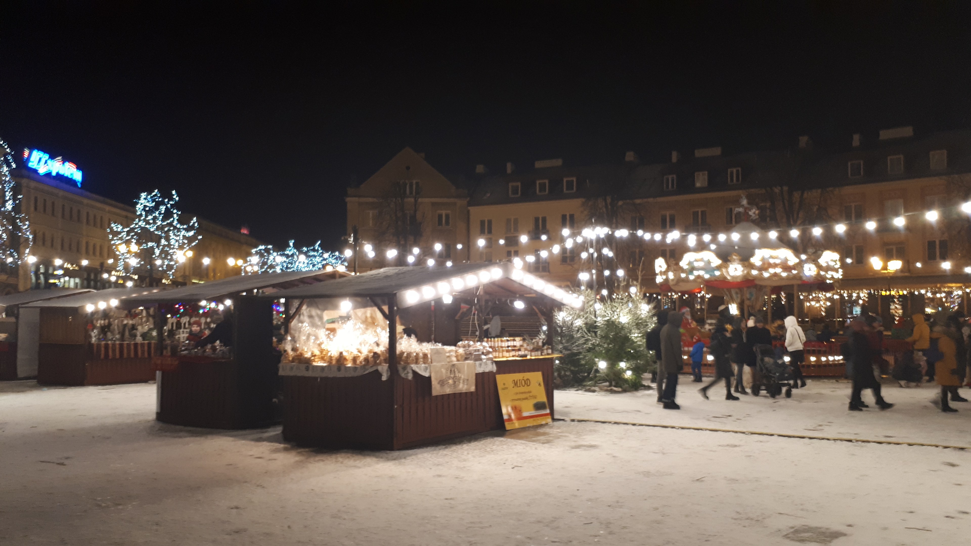 Lebendiger Weihnachtsmarkt an einem verschneiten Abend mit Menschen, Buden, Pflanzen, Bäumen, Gebäuden und Schildern unter einem bewölkten Himmel.