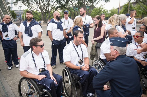 Eine Gruppe von Menschen im Rollstuhl unterhält sich vor einem Hintergrund aus Bäumen, Pfählen, Gebäuden und einem klaren blauen Himmel, wobei einige Mützen und Brillen tragen.