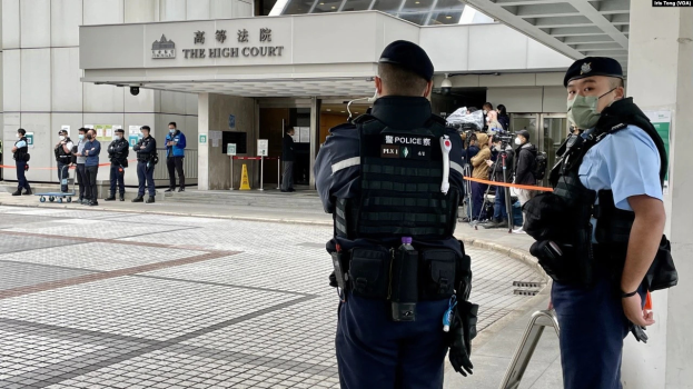 Zwei Polizisten in Uniform, mit Mützen und Masken, stehen vor einem Gebäude am Hong Kong International Airport, umgeben von einer Gruppe von Menschen, einige halten Kameras, mit Stangen mit Bändern im Hintergrund.