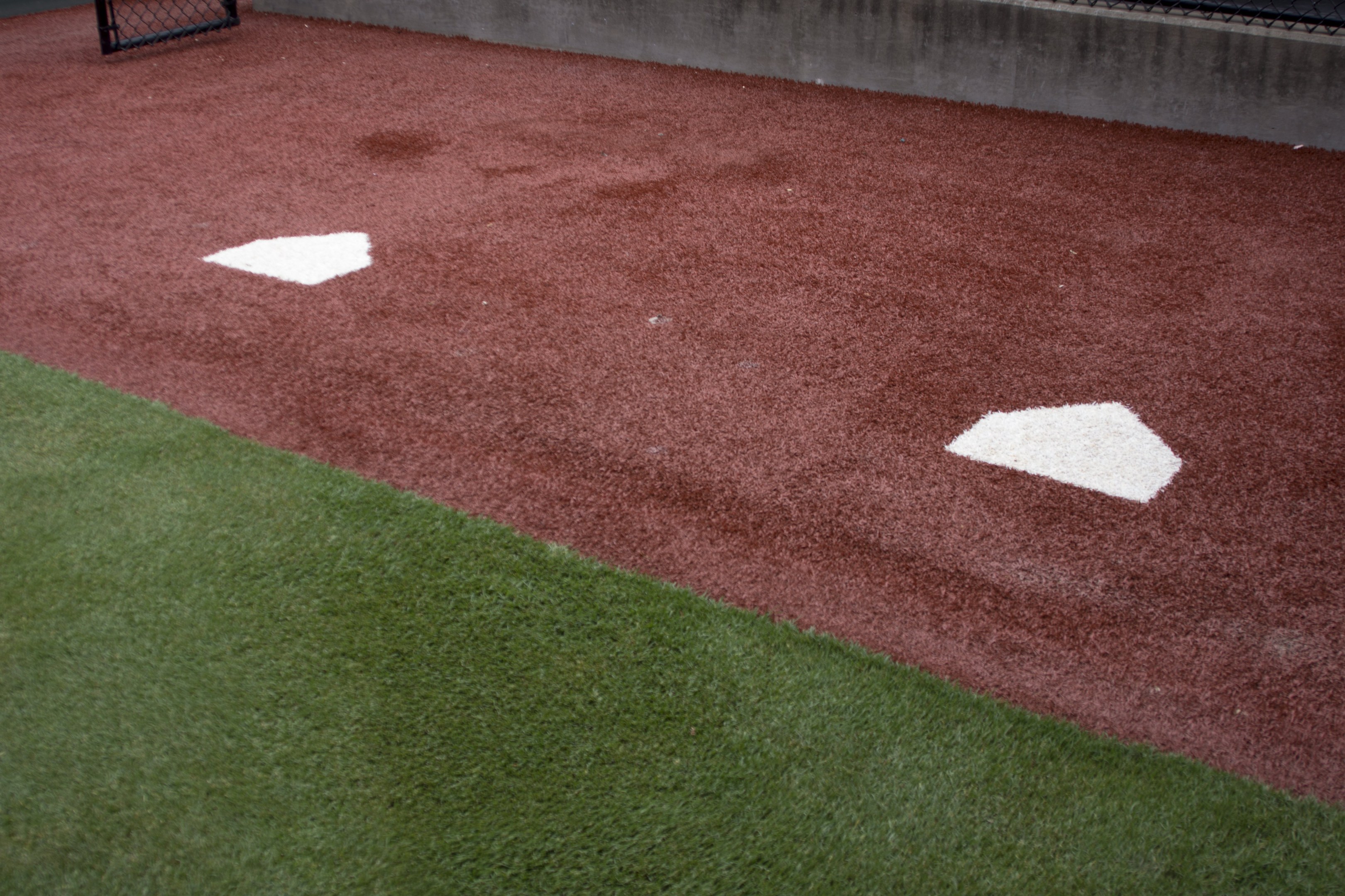 Baseballfeld mit Kunstrasen, umgeben von einem Zaun, mit Home Plate in der Mitte und einer Wand im Hintergrund.