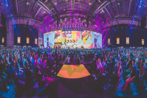 Eine große Menschenmenge, die auf Stühlen vor einer hell erleuchteten Bühne sitzt, mit mehreren stehenden Individuen, wahrscheinlich auf einem Online-Marketing-Rockstars-Event, umgeben von Bildschirmen.