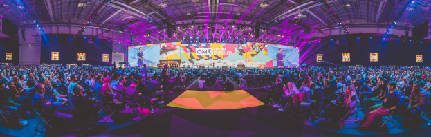 Eine große Menschenmenge, die auf Stühlen vor einer hell erleuchteten Bühne sitzt, mit mehreren stehenden Individuen, wahrscheinlich auf einem Online-Marketing-Rockstars-Event, umgeben von Bildschirmen.