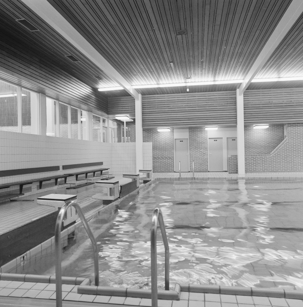 Große Hallenbad mit gefliestem Boden, Metallstötzen, Bänken und klarem Wasser unter sichtbaren Deckenlichtern und Fenstern.