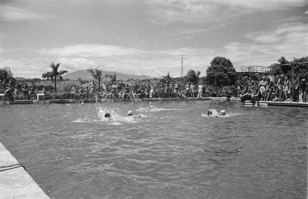 Schwarze und weiße Fotografie von Menschen, die in einem Pool schwimmen, mit Zuschauern hinter einem Zaun, Pfählen, einem Turm, Bäumen, Hügeln und einem bewölkten Himmel.