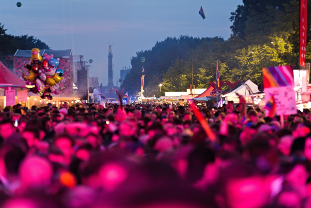 Eine große Menschenmenge auf einem nächtlichen Musikfestival mit farbiger Beleuchtung, umgeben von Zelten, Fahnen und Strukturen, mit Bäumen, einem Turm und einem klaren Himmel im Hintergrund.