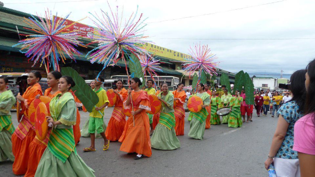 Eine Gruppe von Menschen in bunten Kostümen geht öffentlich eine Straße entlang, mit einer Frau auf der rechten Seite, die eine Flasche hölt, und Fahrzeugen, Gebäuden, Bäumen, Masten, Drähten und Himmel im Hintergrund.