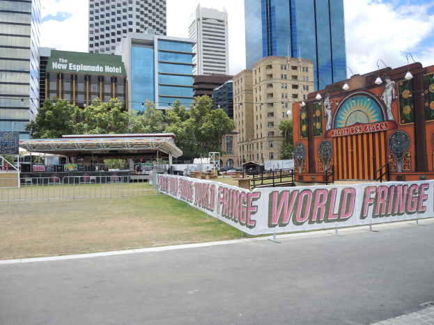 Ein Karnevalsfahrgeschäft ist in einer Stadtstraße aufgebaut, umgeben von hohen Gebäuden, Bäumen und einem bewölkten Himmel, mit einer Straße, einem Geländer und einer Fahne mit Text im Vordergrund.