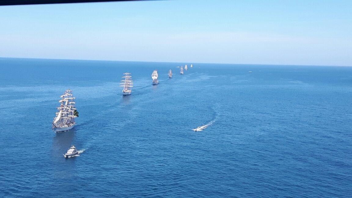 Eine große Flotte von Segelschiffen, die in Formation auf einem sonnigen Tag mit klarem blauem Himmel und ruhiger See segeln.