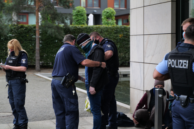 Eine Gruppe von Polizisten steht vor einem Gebäude, während eine Frau ein Buch und einen Stift hält, mit Menschen in der Nähe und Grün im Hintergrund.