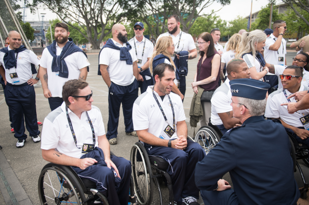 Eine Gruppe von Menschen in Rollstühlen unterhält sich vor einer Kulisse mit Bäumen, Pfählen, Gebäuden und einem klaren blauen Himmel, wobei einige Mützen und Brillen tragen, was auf eine Teilnahme an den Paralympischen Spielen hindeutet.