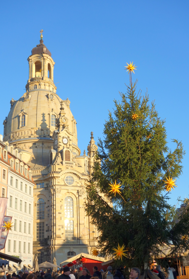 Ein geschäftiger Weihnachtsmarkt in Dresden, Deutschland, mit einem großen Weihnachtsbaum vor einer Kirche, vielen Menschen drumherum und einer Banner mit Text auf der linken Seite, unter einem sichtbaren Himmel.