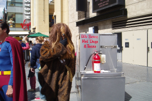 Eine Menschenmenge, darunter einige in Kostümen, versammelte sich vor Gebäuden mit Schildern, einer Box mit Schrift und einem Feuerlöscher.