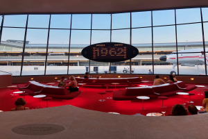 Eine Gruppe von Menschen sitzt in einem geräumigen Raum mit roten Sofas und Tischen, Glaswände zeigen ein Flugzeug, Fahrzeuge, Gebäude, Pfosten und den Himmel, mit einer Tafel, die darauf hinweist, dass es die TWA Lounge am Frankfurt Airport ist.