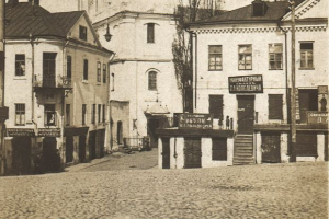 Ein altes Schwarz-Weiß-Foto einer Stadtstraße mit Gebäuden, Fenstern, Türen, Stufen, Geländern, Texttafeln, Laternen, Bäumen und einer Straße unten.
