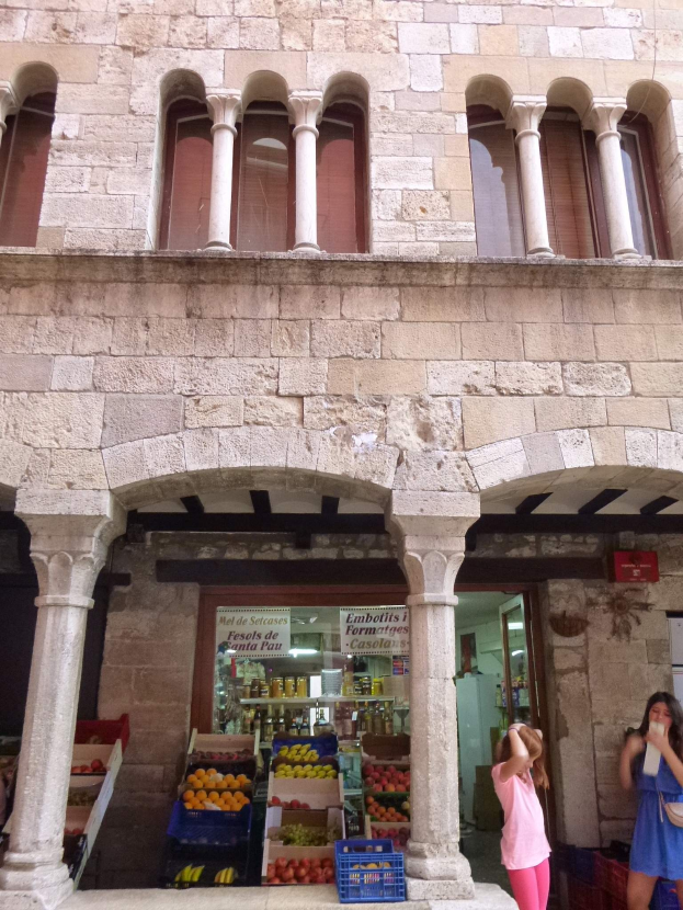 Zwei Frauen stehen vor einem Gebäude mit Säulen, Fenstern und einer Tür, das wie ein Obst- und Gemüsemarkt mit Körben voller Obst und einer Tafel mit Text darin aussieht.