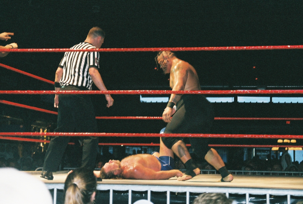 Zwei Boxer stehen im Ring mit roten Seilen, während ein Mann in einem weißen und schwarzen gestreiften T-Shirt von der Seite zusieht.