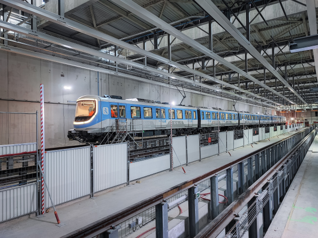 Blauer und weißer Zug fährt durch eine große Indoor-Station mit einer Plattform auf der rechten Seite, einer Wand im Hintergrund und einer Decke, die mit Stangen und Lampen ausgestattet ist.