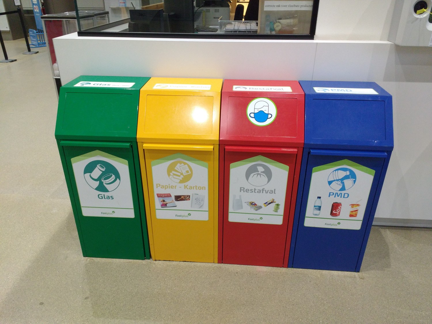 Vier farbenfrohe Mülltonnen in einer Reihe neben einem Tresen mit einem Monitor und anderen Gegenständen, mit einer Person auf der linken Seite und an der Wand befestigten Gegenständen auf der rechten Seite.