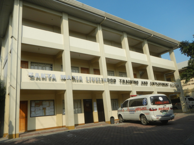 Außenansicht des Santa Maria Lebensmittel-Trainings- und Arbeitsplatz-Zentrums mit Fenstern, Türen und Beschriftung, umgeben von Bäumen mit Fahrzeugen auf der Straße und einem klaren blauen Himmel.
