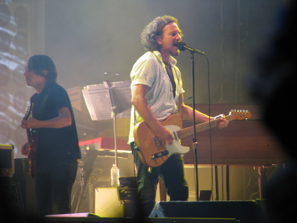 Ein Mann in einem weißen Hemd spielt Gitarre und singt in ein Mikrofon auf der Bühne, neben ihm ein anderer Mann in einem schwarzen Hemd, der ebenfalls Gitarre spielt; ein Piano, Kabel, Lautsprecher und Lichter sind im Hintergrund zu sehen.