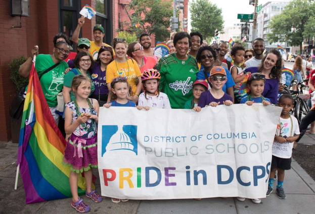Gruppe von Menschen mit einem "District of Columbia Public Schools Pride in DCPS"-Schild, Kinder mit einer Fahne, ein Radfahrer, Bäume, Gebäude, Straßenschilder und ein bewölkter Himmel.