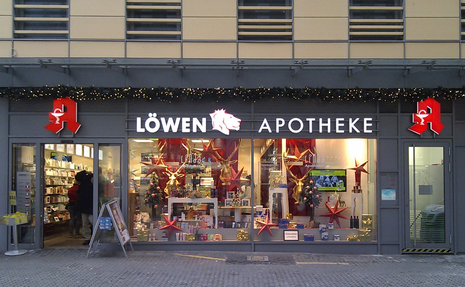Ein Schaufenster mit einem Schild, auf dem "Löwen Apotheke" steht, und einer Straße unten, mit Regalen voller verschiedener Gegenstände und ein paar Menschen, die drinnen stöbern.