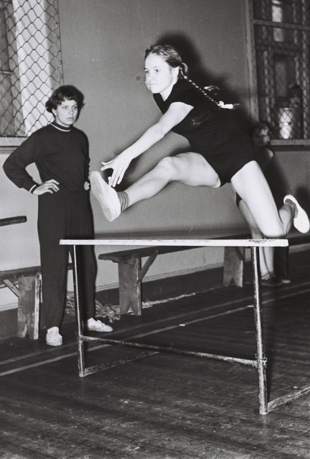 Ein Schwarz-Weiß-Foto einer Frau in einem Einteiler, die über ein Hindernis in einer Turnhalle springt und die Arme ausstreckt, während im Hintergrund eine Person steht und eine Wand, ein Fenster und ein Netz zu sehen sind.