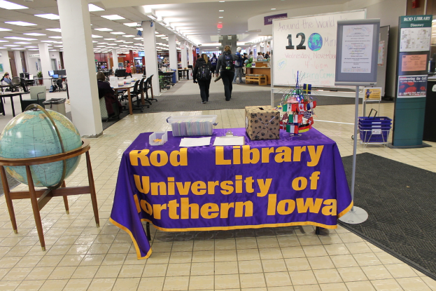 Eine Ausstellung mit einem Tisch, auf dem verschiedene Landesflaggen, eine Kiste, Papiere und eine Fahne sowie links daneben eine Weltkugel, rechts daneben eine Anschlagtafel mit einigen Menschen in der Nähe und mehrere Tische und Stühle im Hintergrund zu sehen sind.